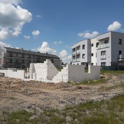 Firma Remontowo budowlana Łukasz Beyer - Budowa domu z białych bloczków na działce, w tle budynek w rusztowaniach i nowoczesny apartamentowiec pod błękitnym niebem.