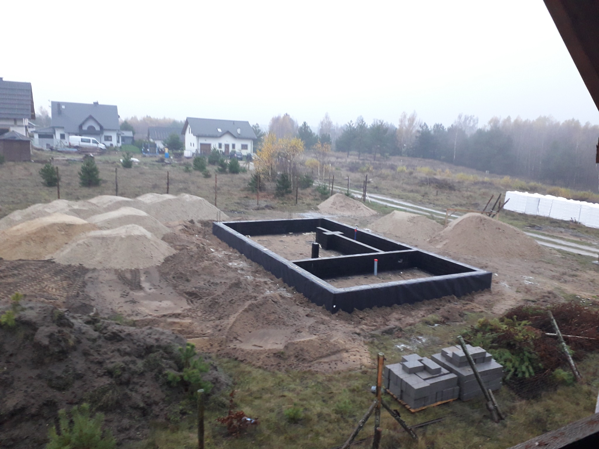 Fundament domu w trakcie budowy, zabezpieczony czarną folią, widoczne sterty piasku i bloczki betonowe na działce w jesiennej scenerii z domami w tle.