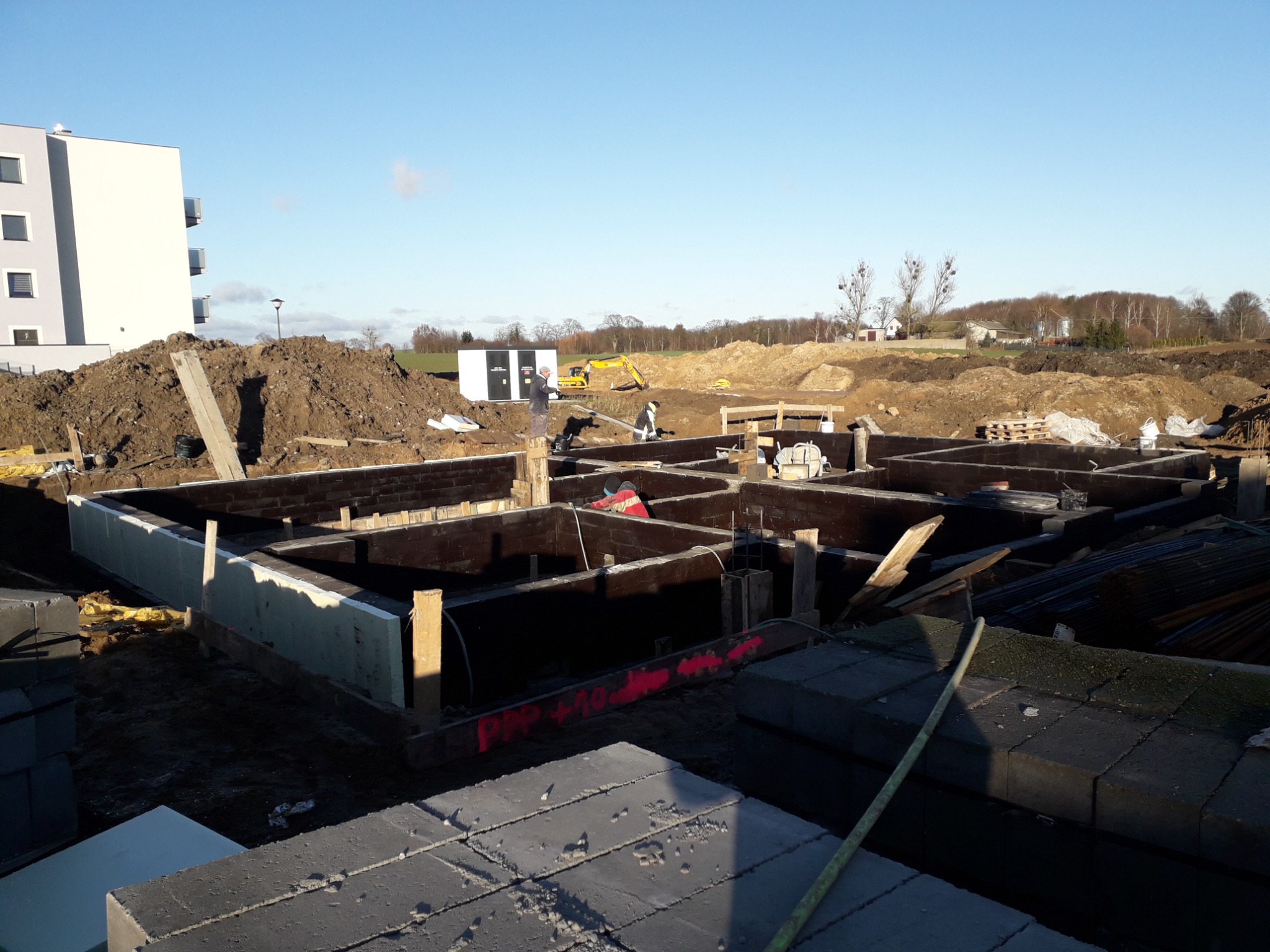 Wykop pod fundamenty domu murowanego z deskowaniem i izolacją termiczną, widoczny pracownik w czerwonej kurtce, w tle budynek wielorodzinny i koparka.