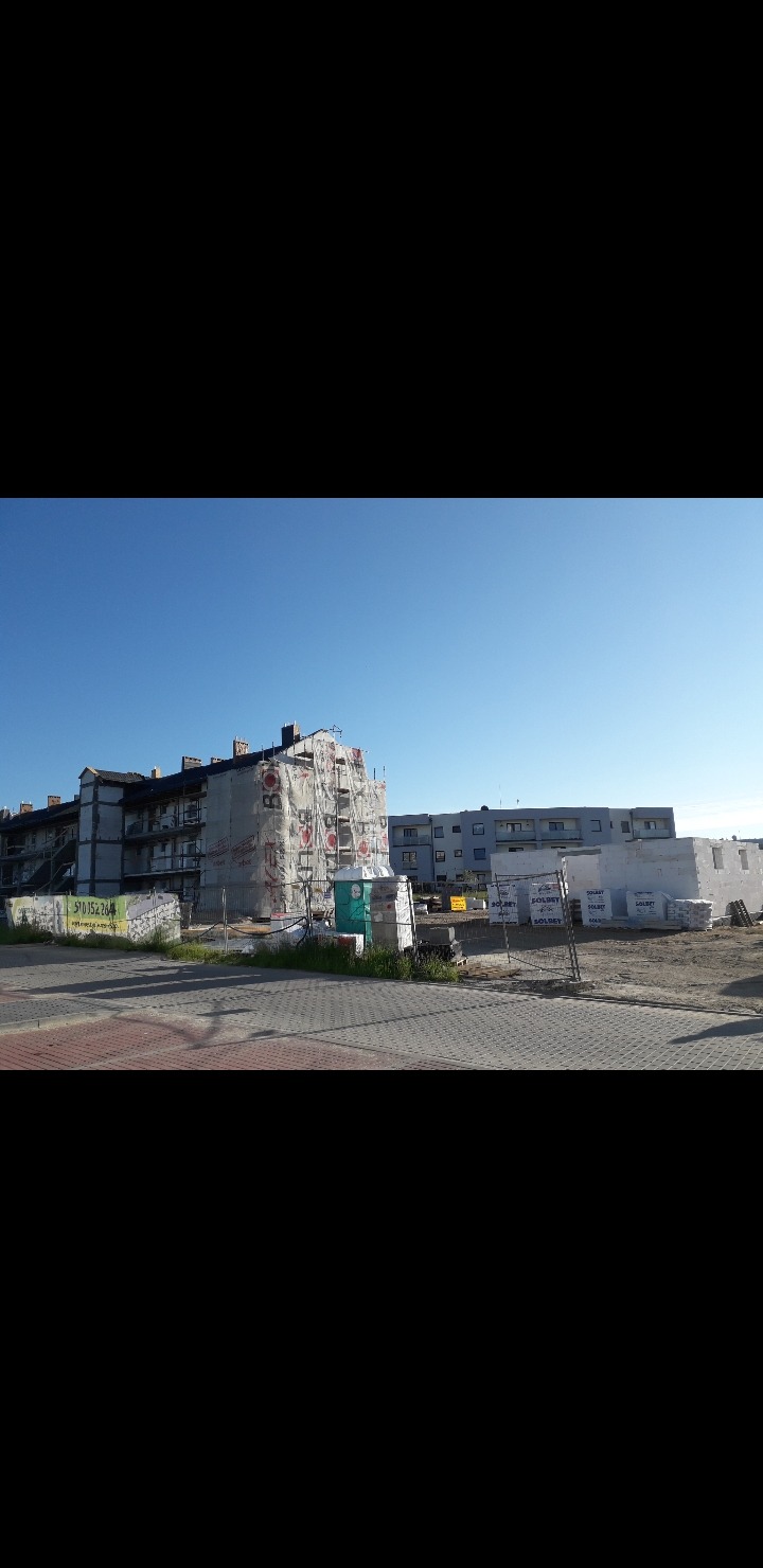 Budynek wielorodzinny w trakcie prac budowlanych, osłonięty siatką rusztowaniową z reklamą, widoczne materiały budowlane i przenośna toaleta.