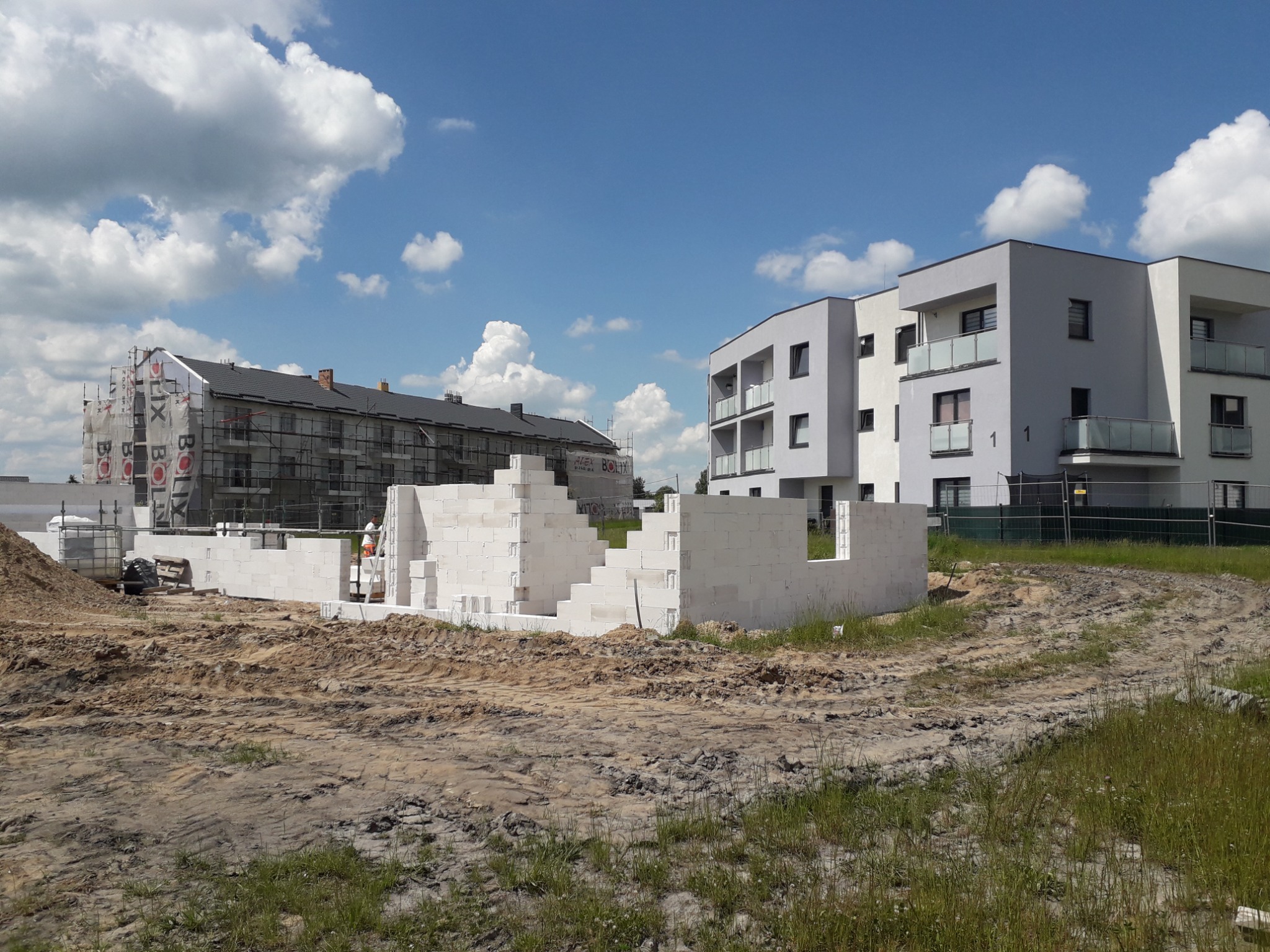 Budowa domu z białych bloczków na działce, w tle budynek w rusztowaniach i nowoczesny apartamentowiec pod błękitnym niebem.