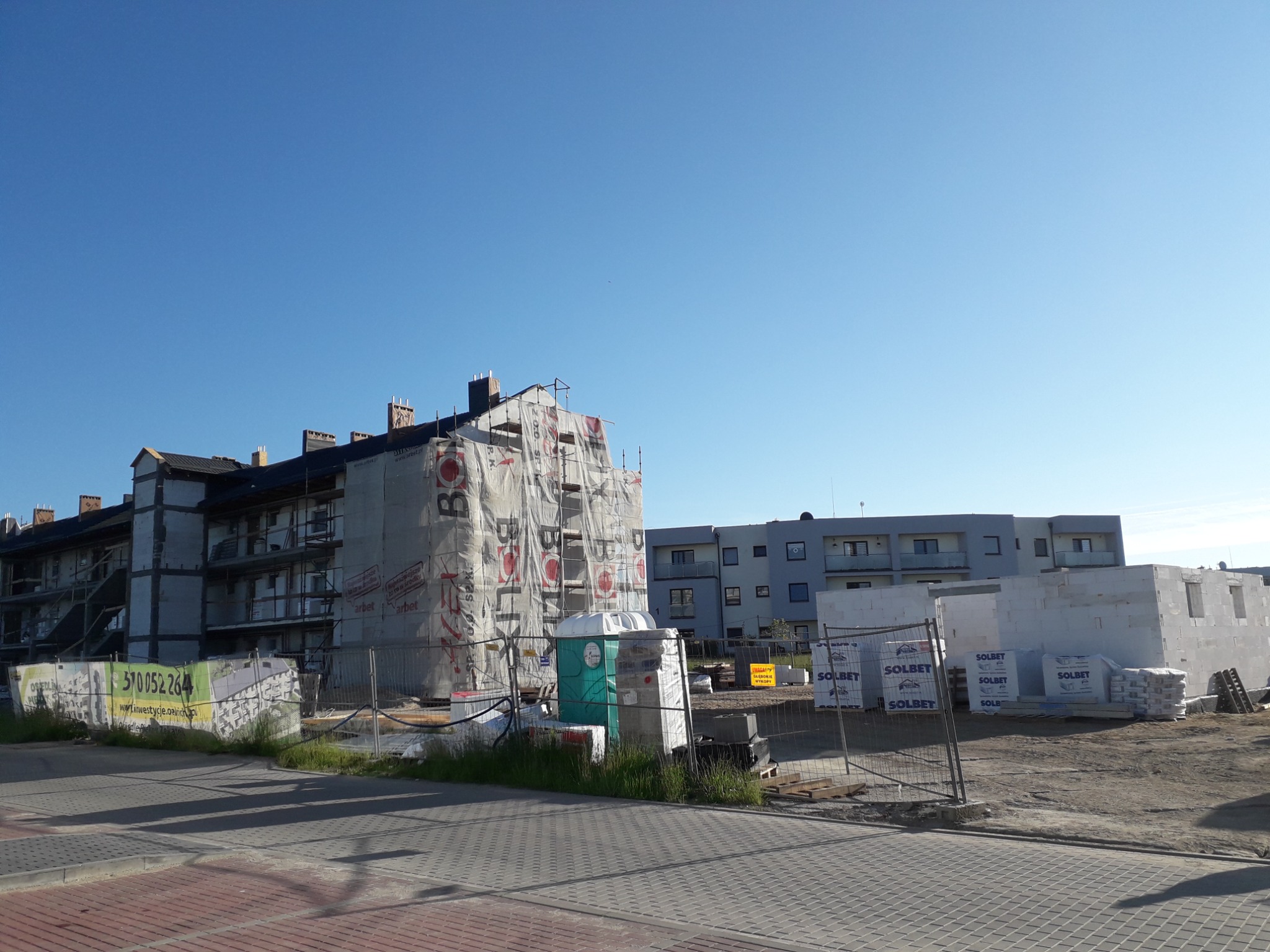 Budowa wielorodzinnego budynku z pustaków Solbet, zabezpieczonego siatką ochronną z reklamą, widok w słoneczny dzień.