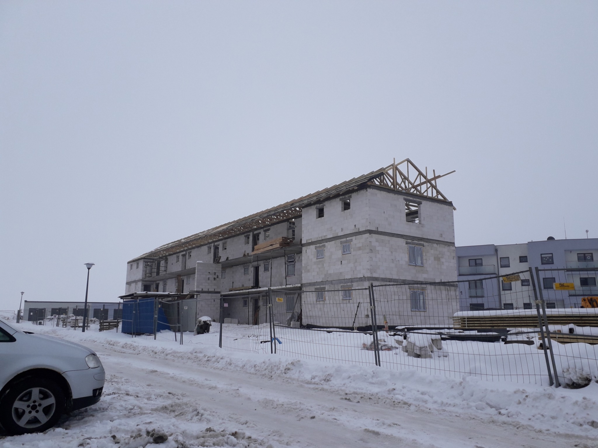 Budynek wielorodzinny w stanie surowym otwartym, z widoczną konstrukcją dachu, wykonany z białych bloczków, otoczony śniegiem i ogrodzeniem budowlanym, z zaparkowanym samochodem osobowym w pobliżu.