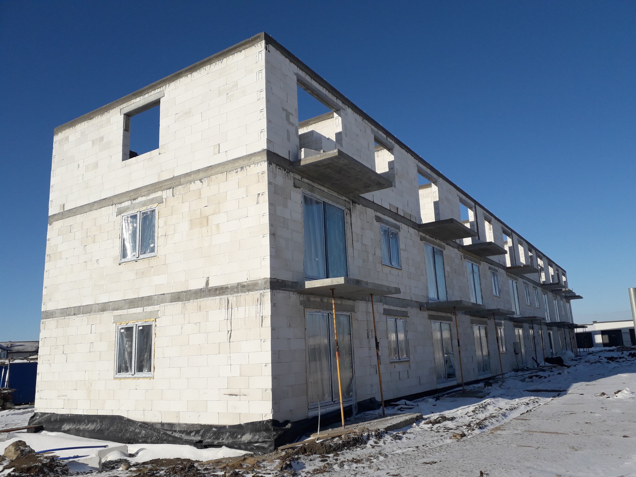 Wielopiętrowy budynek w stanie surowym, wzniesiony z białych bloczków, z widocznymi oknami zabezpieczonymi folią i fragmentami rusztowań, na tle błękitnego nieba i zimowego krajobrazu.
