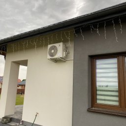 Defrost Dawid Krawiec - Zewnętrzny klimatyzator marki Gree zamontowany na elewacji domu, ozdobiony świątecznymi lampkami, widoczny kran i wąż ogrodowy.