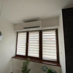 Defrost Dawid Krawiec - Biały klimatyzator ścienny zamontowany nad oknem z roletami dzień-noc, widok z dołu, z doniczkowymi roślinami w tle.