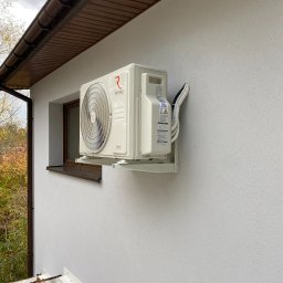 Defrost Dawid Krawiec - Zewnętrzna jednostka klimatyzacji Rotenso Hiro Series zamontowana na jasnej ścianie budynku, widoczne okno obok i brązowe elementy dachu.