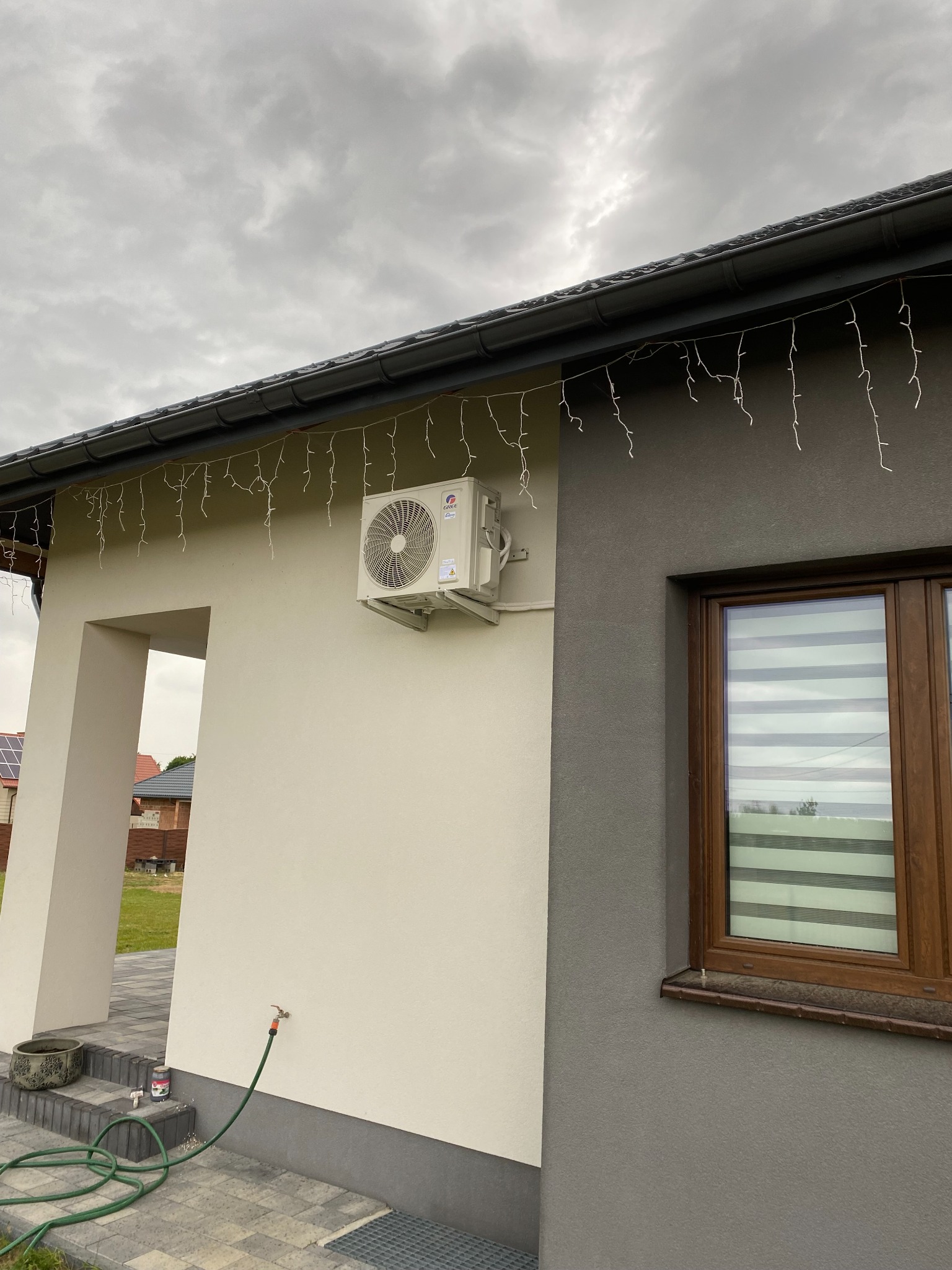 Zewnętrzny klimatyzator marki Gree zamontowany na elewacji domu, ozdobiony świątecznymi lampkami, widoczny kran i wąż ogrodowy.