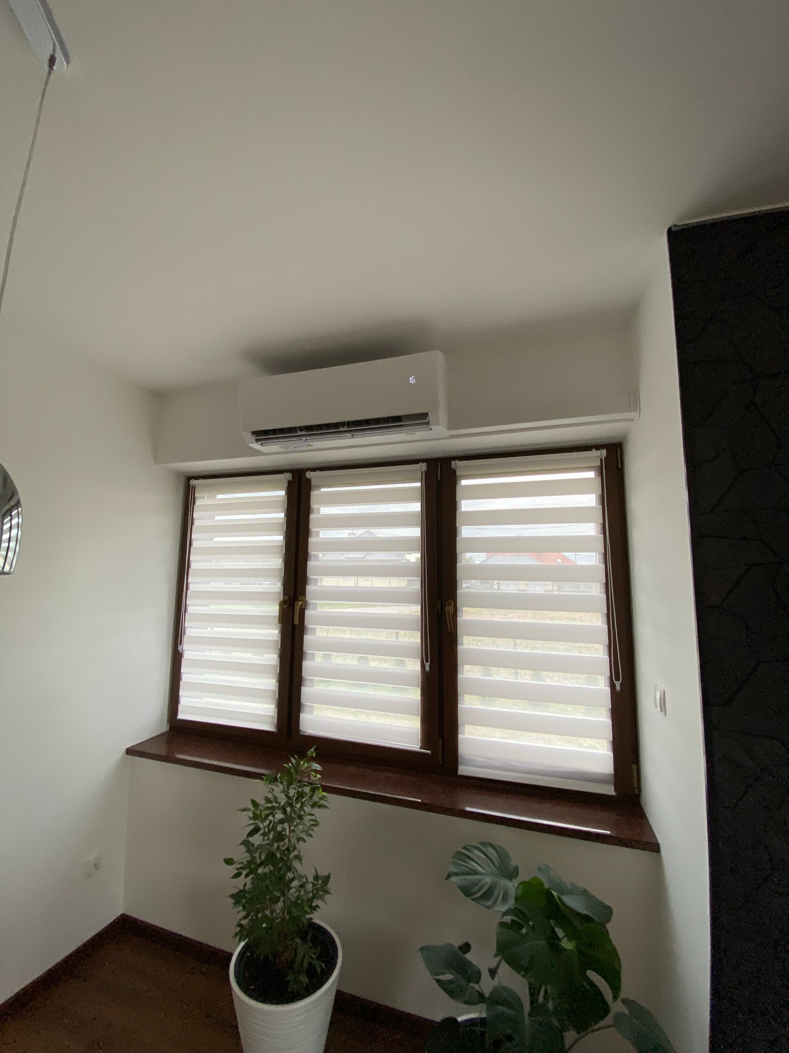 Biały klimatyzator ścienny zamontowany nad oknem z roletami dzień-noc, widok z dołu, z doniczkowymi roślinami w tle.