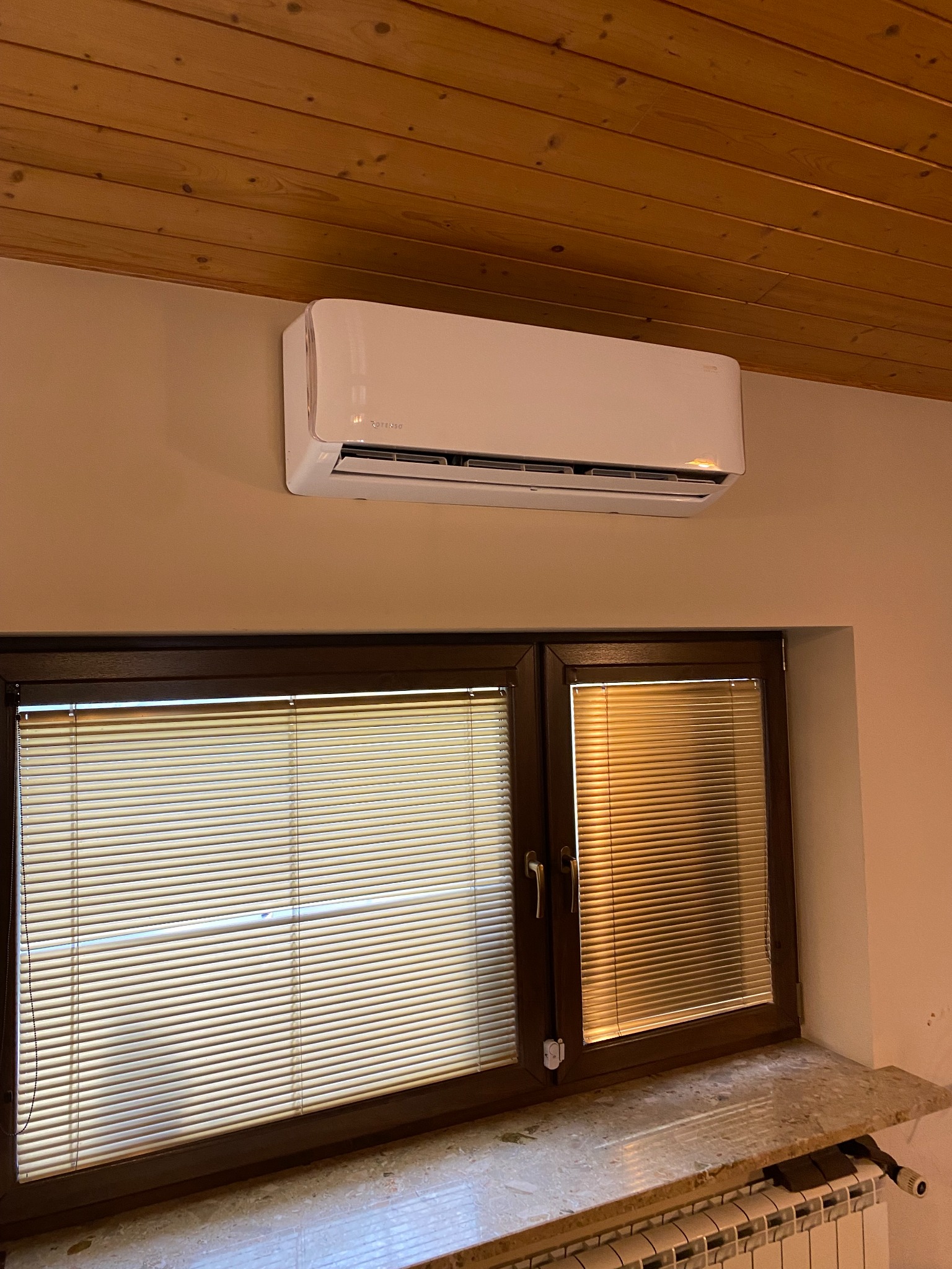 Biały klimatyzator zamontowany nad oknem z żaluzjami w pokoju z drewnianym sufitem.