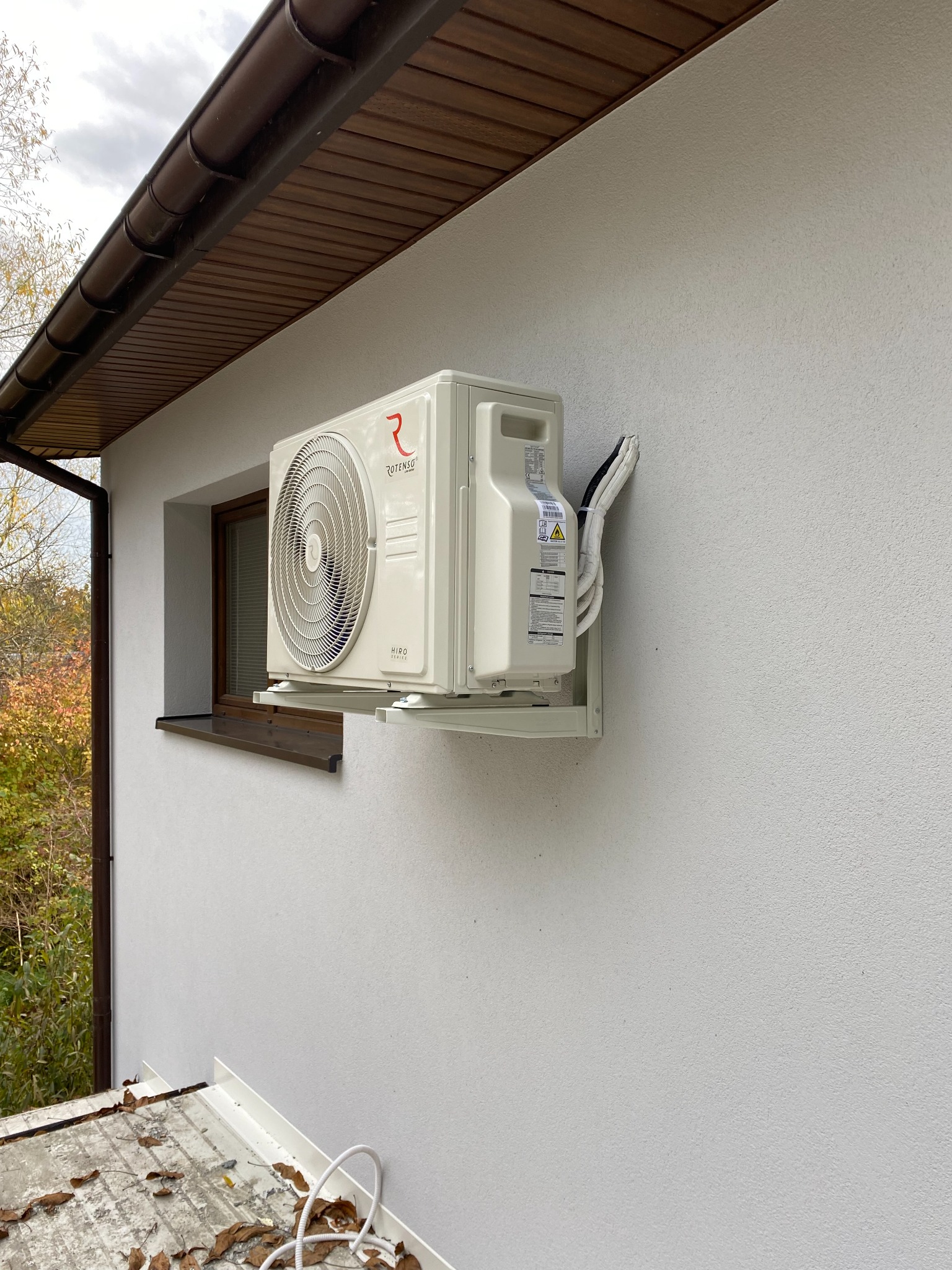 Zewnętrzna jednostka klimatyzacji Rotenso Hiro Series zamontowana na jasnej ścianie budynku, widoczne okno obok i brązowe elementy dachu.