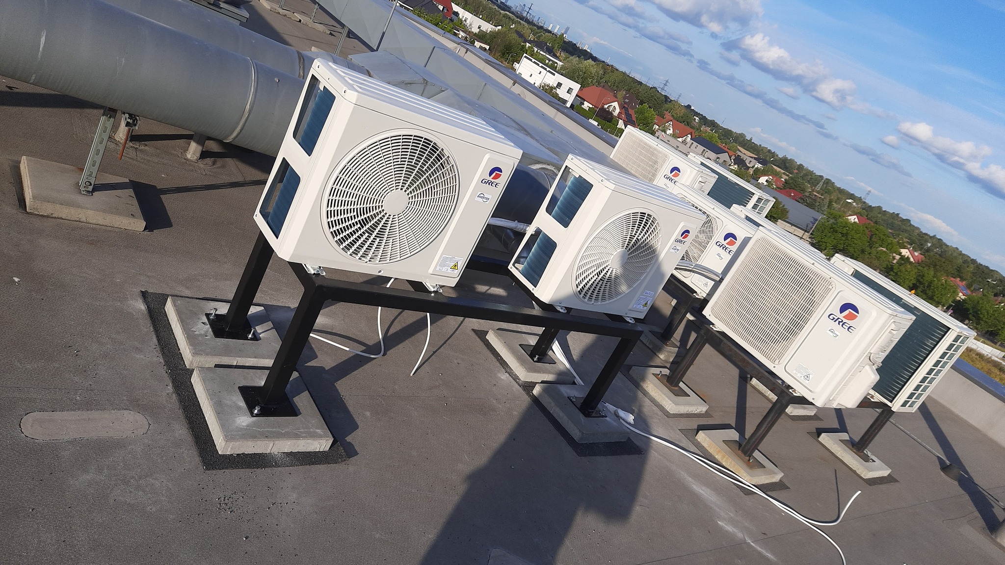 Rząd klimatyzatorów marki Gree zamontowanych na dachu budynku, widoczne metalowe stelaże i betonowe podstawy, w tle zabudowa miejska i błękitne niebo.
