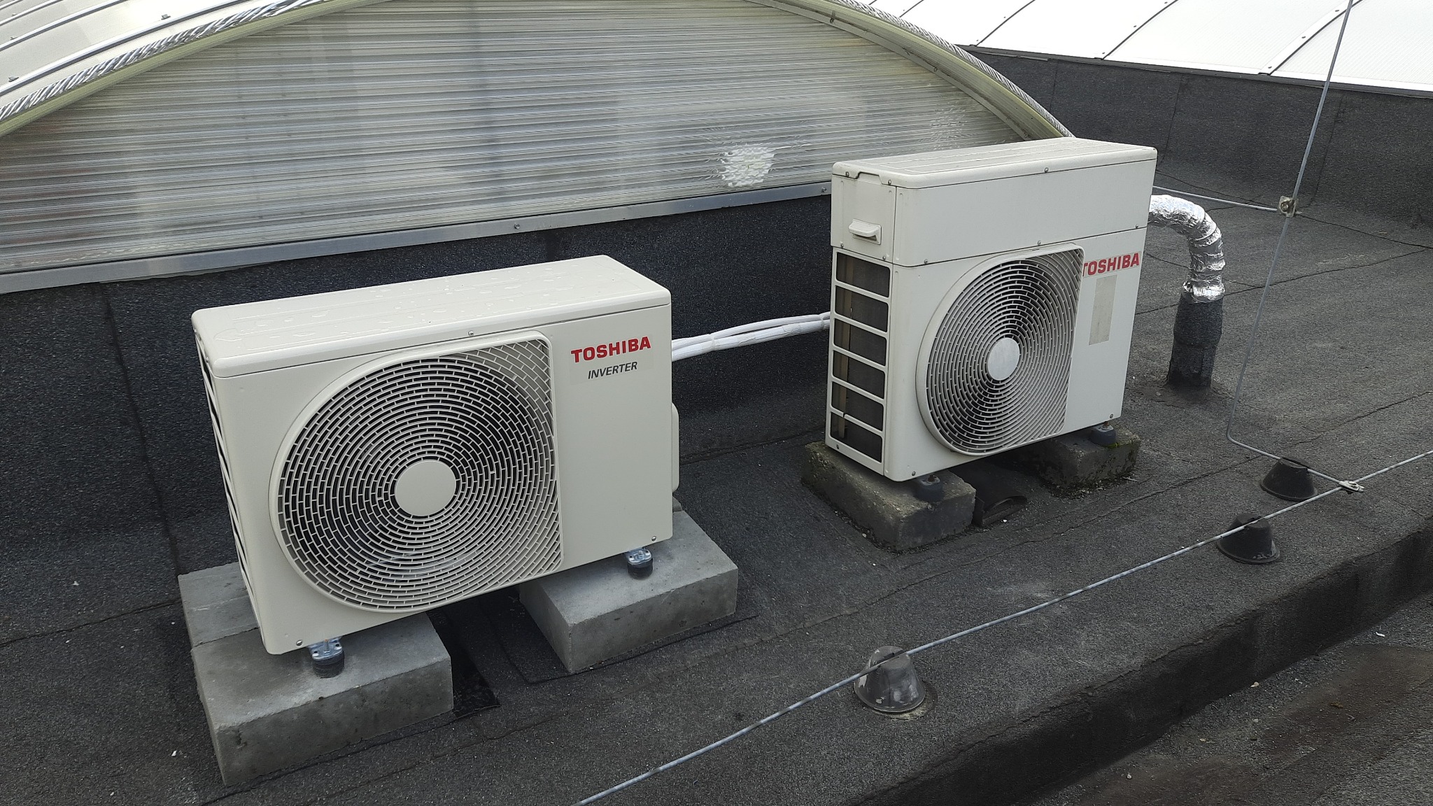 Dwa zewnętrzne agregaty klimatyzacyjne Toshiba Inverter, umieszczone na betonowych podstawach na płaskim dachu pokrytym papą, z widocznymi przewodami i elementami instalacji odgromowej.