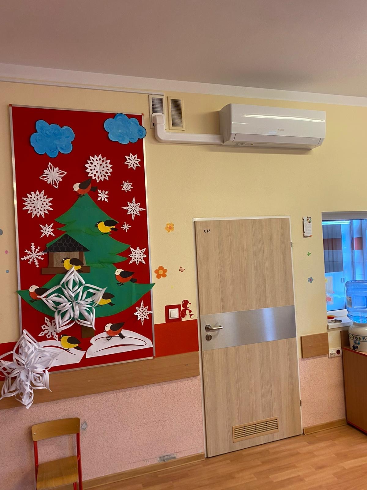 Wnętrze pokoju z klimatyzatorem na ścianie nad drzwiami, obok dekoracja świąteczna z papieru w postaci choinki i płatków śniegu.