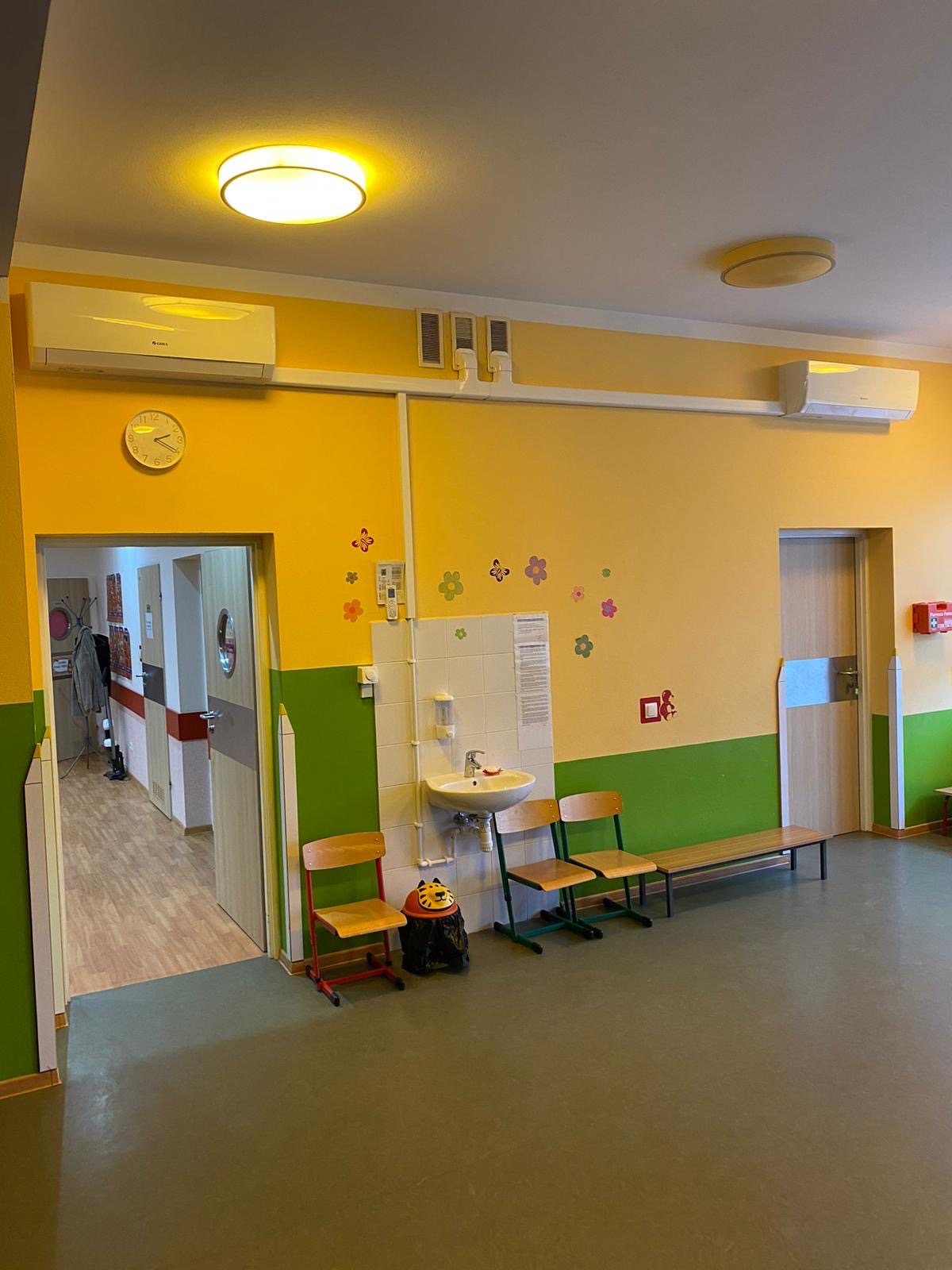 Wnętrze przedszkola z dwoma klimatyzatorami zamontowanymi pod sufitem, umywalką, krzesłami, zegarem ściennym wskazującym godzinę 11:15 i dekoracjami ściennymi w postaci kwiatków i ludzika.
