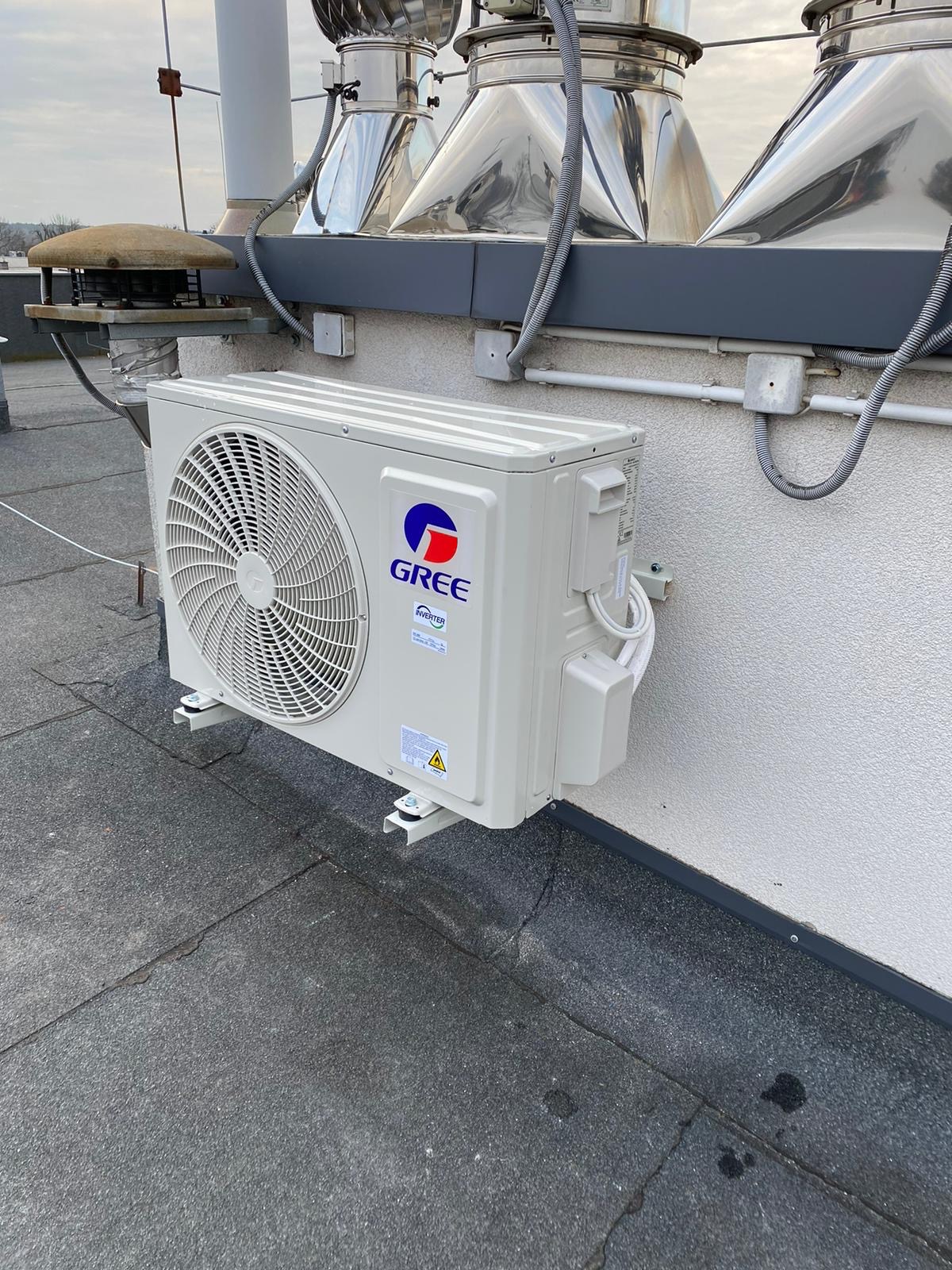 Zewnętrzna jednostka klimatyzacji Gree zamontowana na dachu budynku, widoczne kominy wentylacyjne w tle.