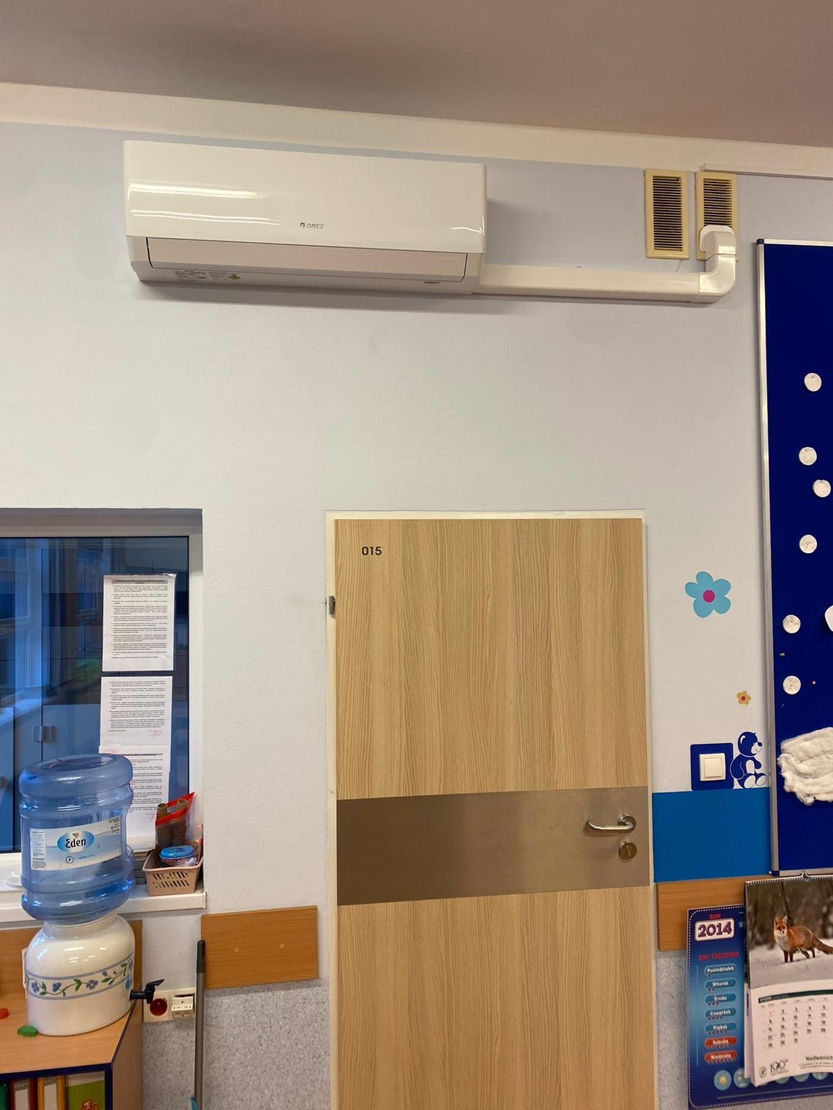 Biała klimatyzacja marki Gree zamontowana na ścianie w pomieszczeniu, z widocznymi przewodami i kratką wentylacyjną, nad drewnianymi drzwiami z numerem 015 i plakatem z kalendarzem.