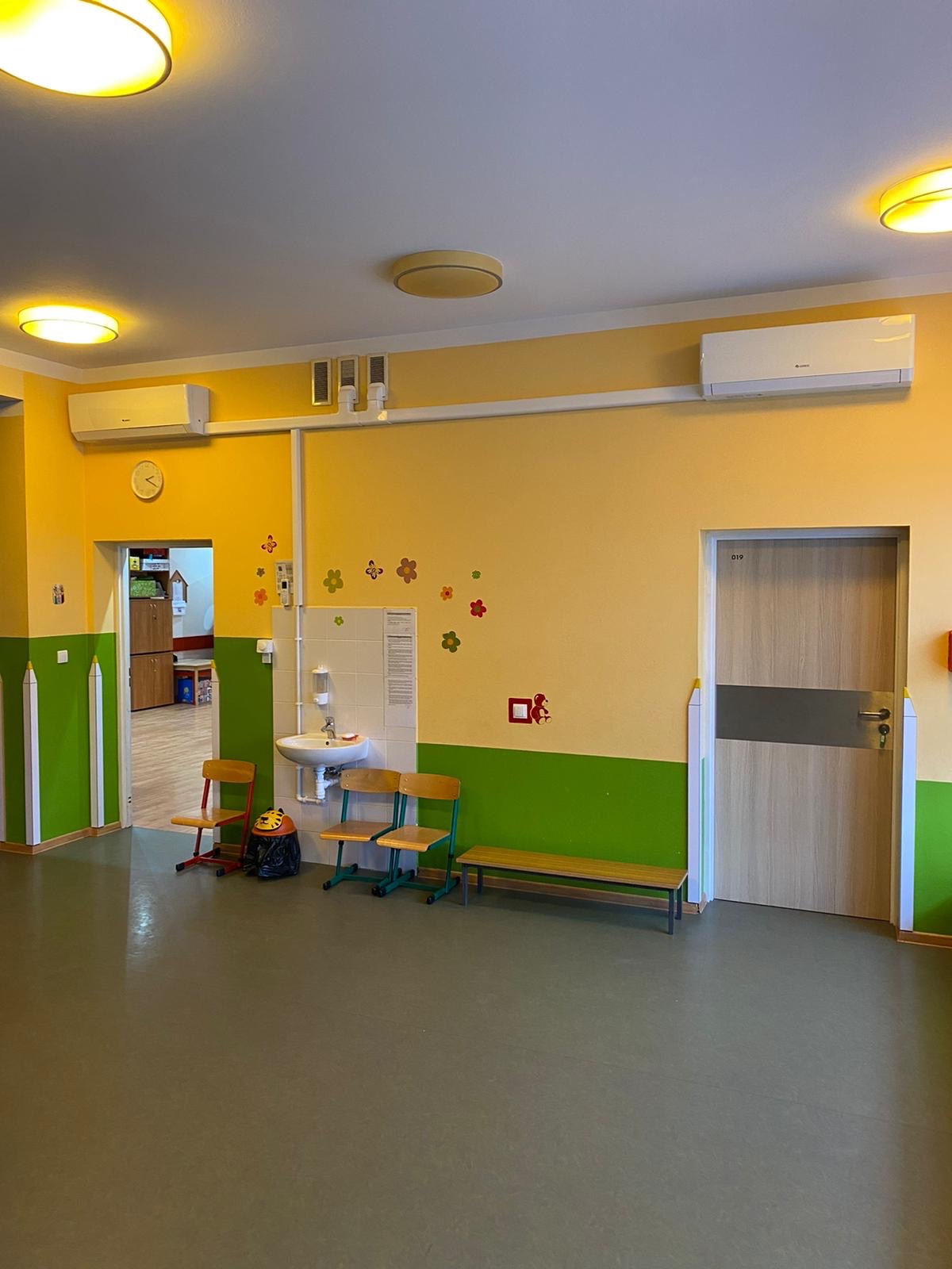 Wnętrze przedszkola z klimatyzatorami na ścianie, umywalką, niskimi krzesłami i dekoracjami ściennymi w postaci kwiatów i kredek.