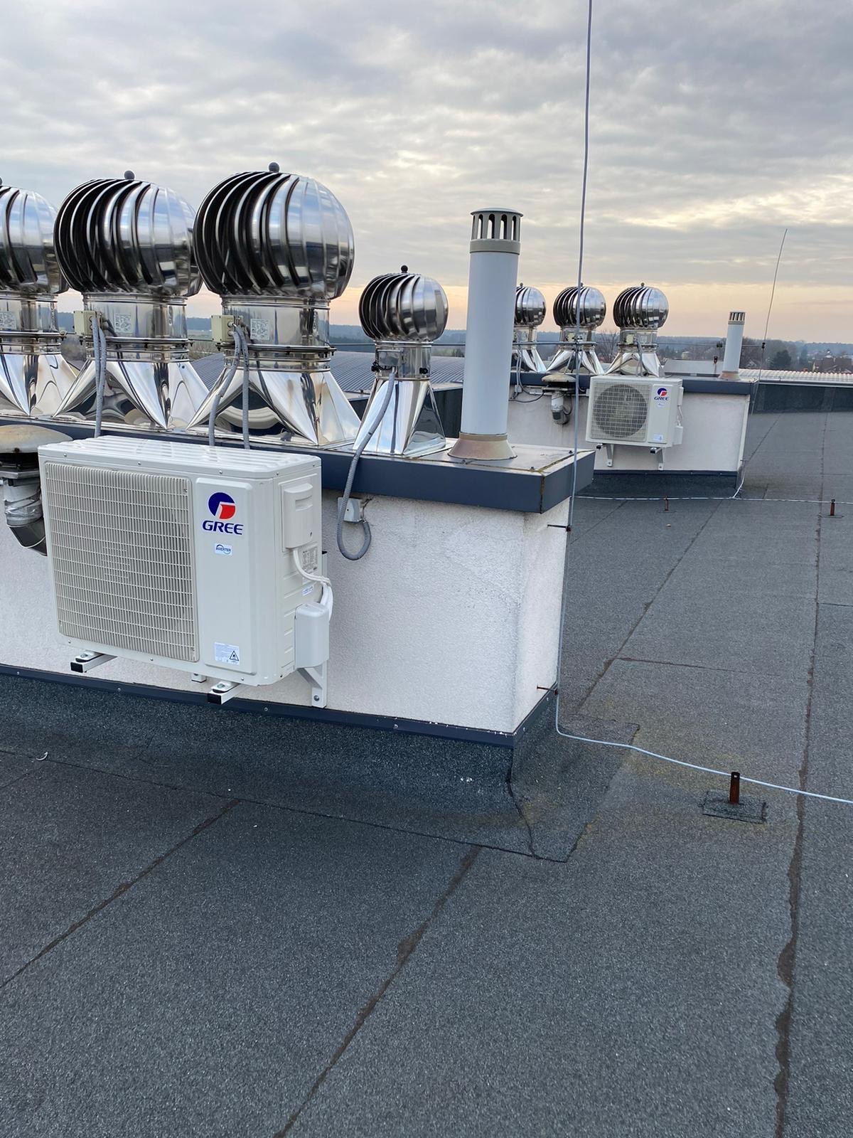 Klimatyzatory marki Gree zamontowane na dachu budynku z licznymi turbowentylatorami i kominami wentylacyjnymi w tle, widok z perspektywy dachu.