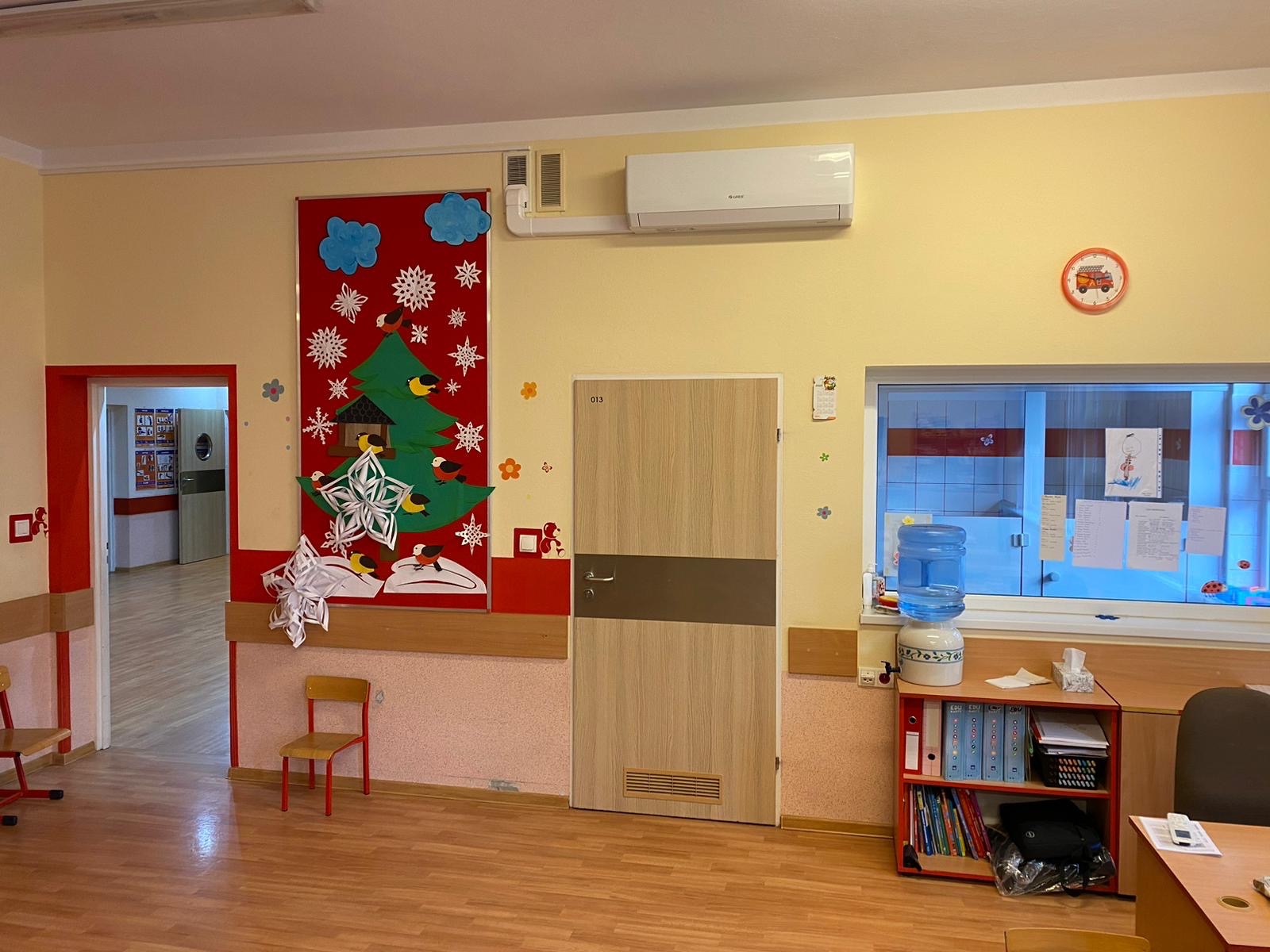 Wnętrze pokoju z klimatyzatorem Gree zamontowanym nad drzwiami, dekoracjami świątecznymi na ścianie i dystrybutorem wody na szafce pod oknem.