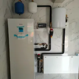 Biała pompa ciepła Panasonic z niebieską taśmą zabezpieczającą ekran, podłączona do białego zasobnika wody i dwóch zbiorników wyrównawczych (niebieski i biały) na tle marmurowych płytek ściennych.