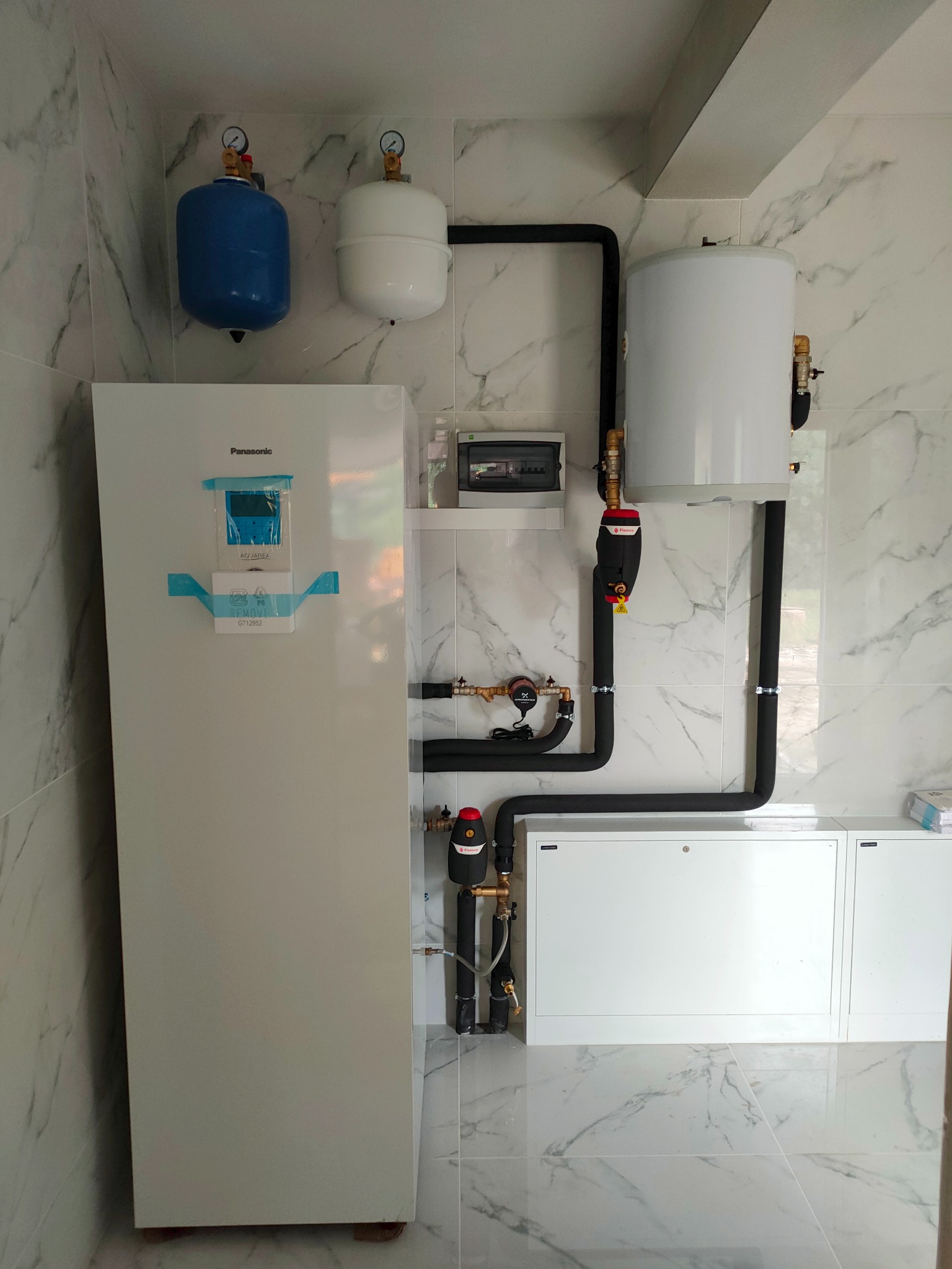 Biała pompa ciepła Panasonic z niebieską taśmą zabezpieczającą ekran, podłączona do białego zasobnika wody i dwóch zbiorników wyrównawczych (niebieski i biały) na tle marmurowych płytek ściennych.
