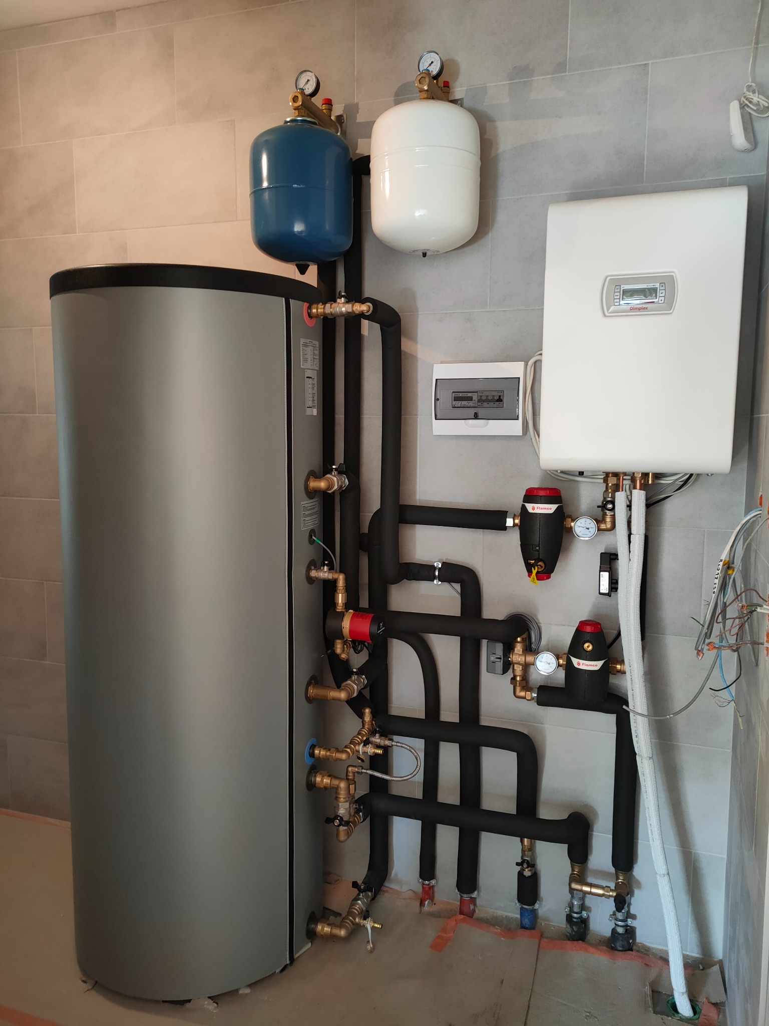 Szary zbiornik buforowy ciepła z podłączonymi rurami w izolacji, dwa naczynia wzbiorcze (białe i niebieskie), biały moduł sterujący Dimplex i rozdzielnica elektryczna na ścianie z szarych płytek.