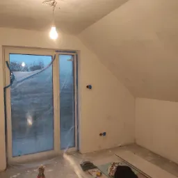 Poddasze w trakcie prac wykończeniowych: przygotowane ściany, zabezpieczone okno folią, widoczne płyty kartonowo-gipsowe, wiadro i klej gipsowy oraz narzędzia.