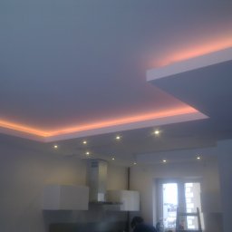 Piotr Kietliński - Nowoczesna kuchnia z podwieszanym sufitem i oświetleniem LED w ciepłym odcieniu, widoczne szafki wiszące i okap, w tle okno z widokiem na zimowy krajobraz.