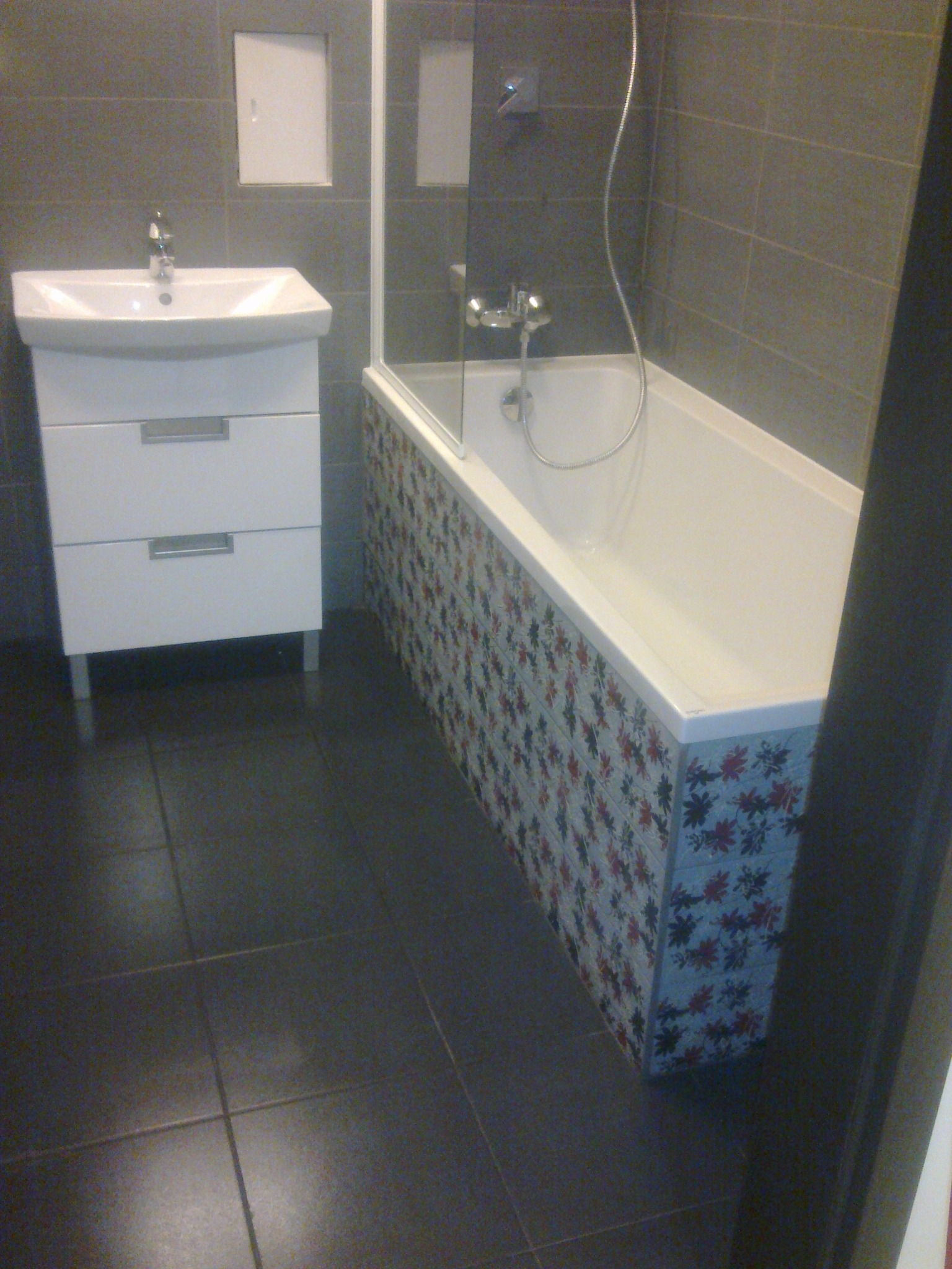 Nowoczesna łazienka z ciemną podłogą, białą umywalką z szafką i wanną z kolorową mozaiką, oddzieloną szklanym parawanem.