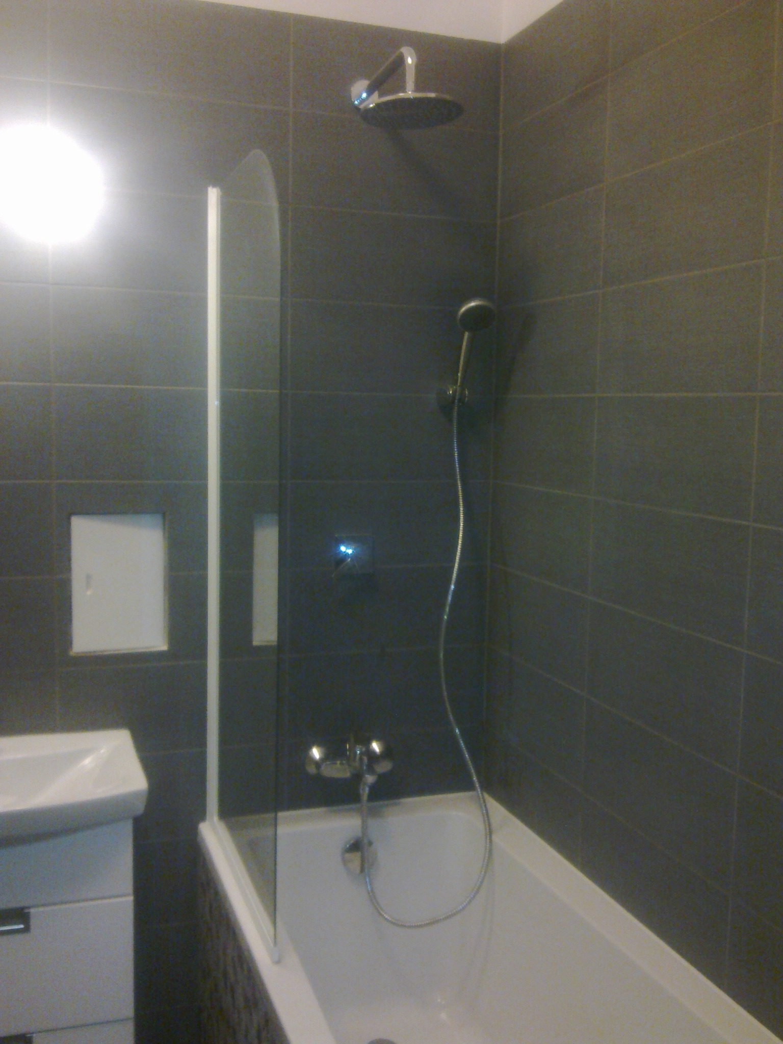 Nowoczesna łazienka z szarymi płytkami, kabiną nawannową z przezroczystego szkła, białą wanną i umywalką z szafką. Widoczny panel dostępowy ukryty w płytkach.