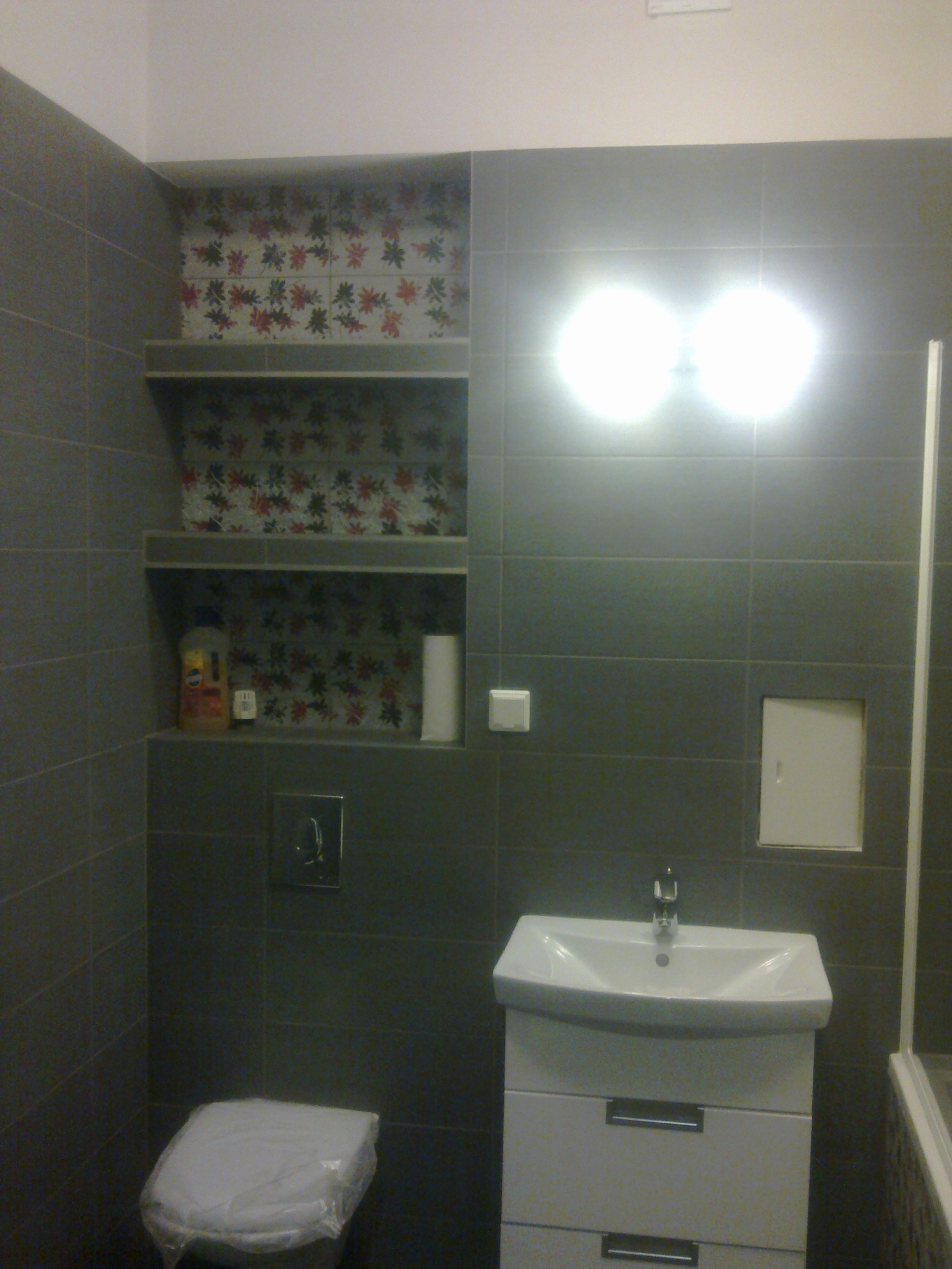 Wykończona łazienka z szarymi płytkami, białą umywalką z szafką, toaletą z zabezpieczeniem i półkami z dekoracyjnymi płytkami w kwiaty.