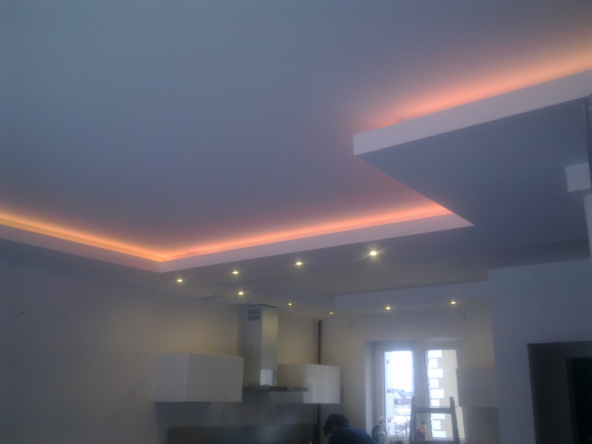 Nowoczesna kuchnia z podwieszanym sufitem i oświetleniem LED w ciepłym odcieniu, widoczne szafki wiszące i okap, w tle okno z widokiem na zimowy krajobraz.