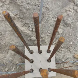 Widok z góry na wystające pręty zbrojeniowe z otworami w kartonowej płycie, umieszczone w ziemi na budowie.