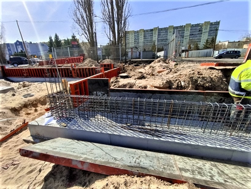 Zbrojenie fundamentu w wykopie z deskowaniem, widoczny fragment pracownika w odblaskowej kamizelce i blok mieszkalny w tle.