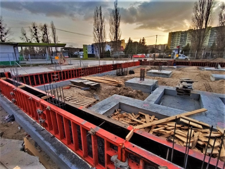 Wykop pod fundamenty z czerwonym szalunkiem i wystającymi prętami zbrojeniowymi, widoczne betonowe stopy fundamentowe, w tle budynki mieszkalne i zachodzące słońce.