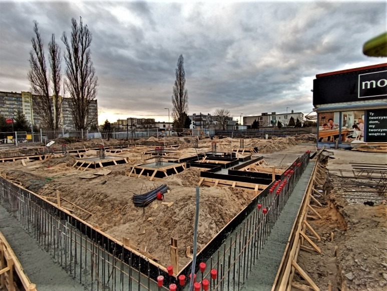 Wykop pod fundamenty z częściowo zalanymi betonem szalunkami i wystającymi prętami zbrojeniowymi, widok na plac budowy z budynkami w tle oraz pochmurnym niebem.