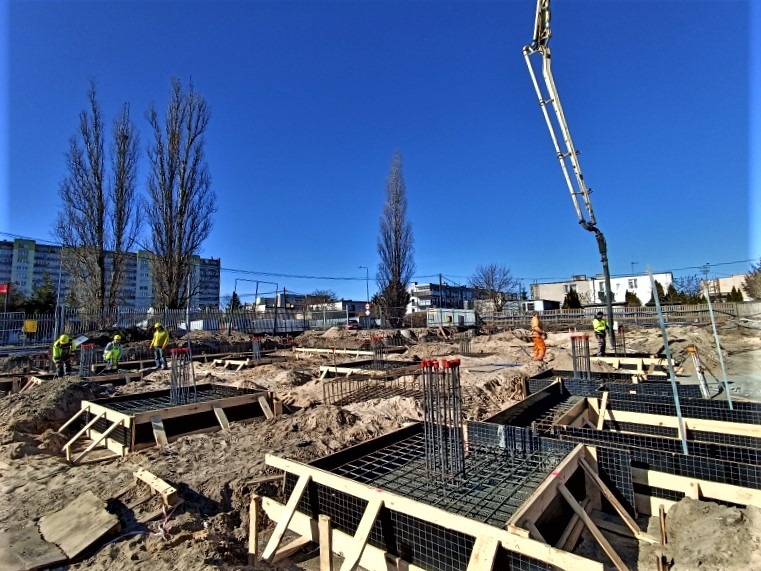 Przygotowanie fundamentów: zbrojenie i szalunki na placu budowy, widoczna pompa do betonu i pracownicy w odblaskowych ubraniach, w tle budynki mieszkalne.