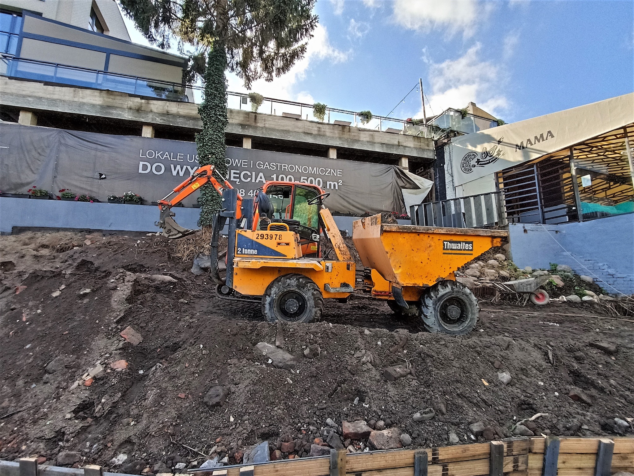 Żółta wywrotka Thwaites i mała koparka na placu budowy, z widocznymi fundamentami i konstrukcją budynku w tle, w tym baner reklamowy lokali użytkowych i restauracji.