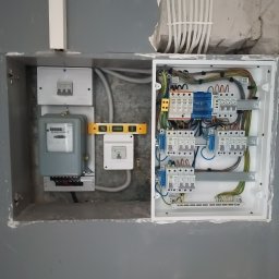 MP Elektro Mikołaj Przybylski - Szara skrzynka z bezpiecznikami i licznikiem elektrycznym, zamontowana w ścianie, widoczny poziomica, przewody w peszlu i instalacja elektryczna w ścianie.
