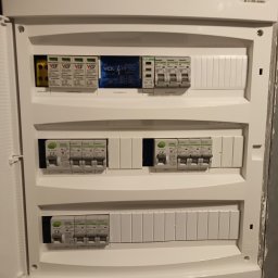 MP Elektro Mikołaj Przybylski - Otwarta skrzynka rozdzielcza z zabezpieczeniami różnicowoprądowymi i ogranicznikami przepięć marki Noark i VCX, widoczne opisy obwodów: salon, kuchnia, zmywarka.