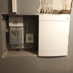 MP Elektro Mikołaj Przybylski - Szara ściana z wnęką, w której umieszczono licznik energii elektrycznej, zabezpieczenie przedlicznikowe i białą skrzynkę rozdzielczą. Nad skrzynką widoczne kable w peszlach.