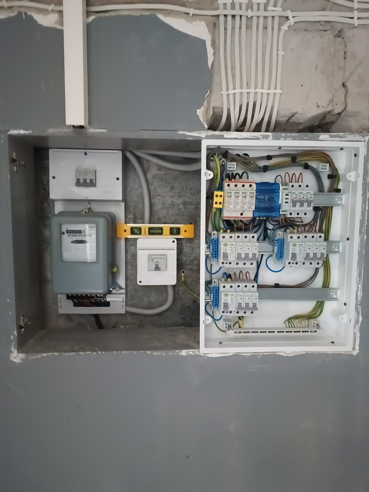 Szara skrzynka z bezpiecznikami i licznikiem elektrycznym, zamontowana w ścianie, widoczny poziomica, przewody w peszlu i instalacja elektryczna w ścianie.