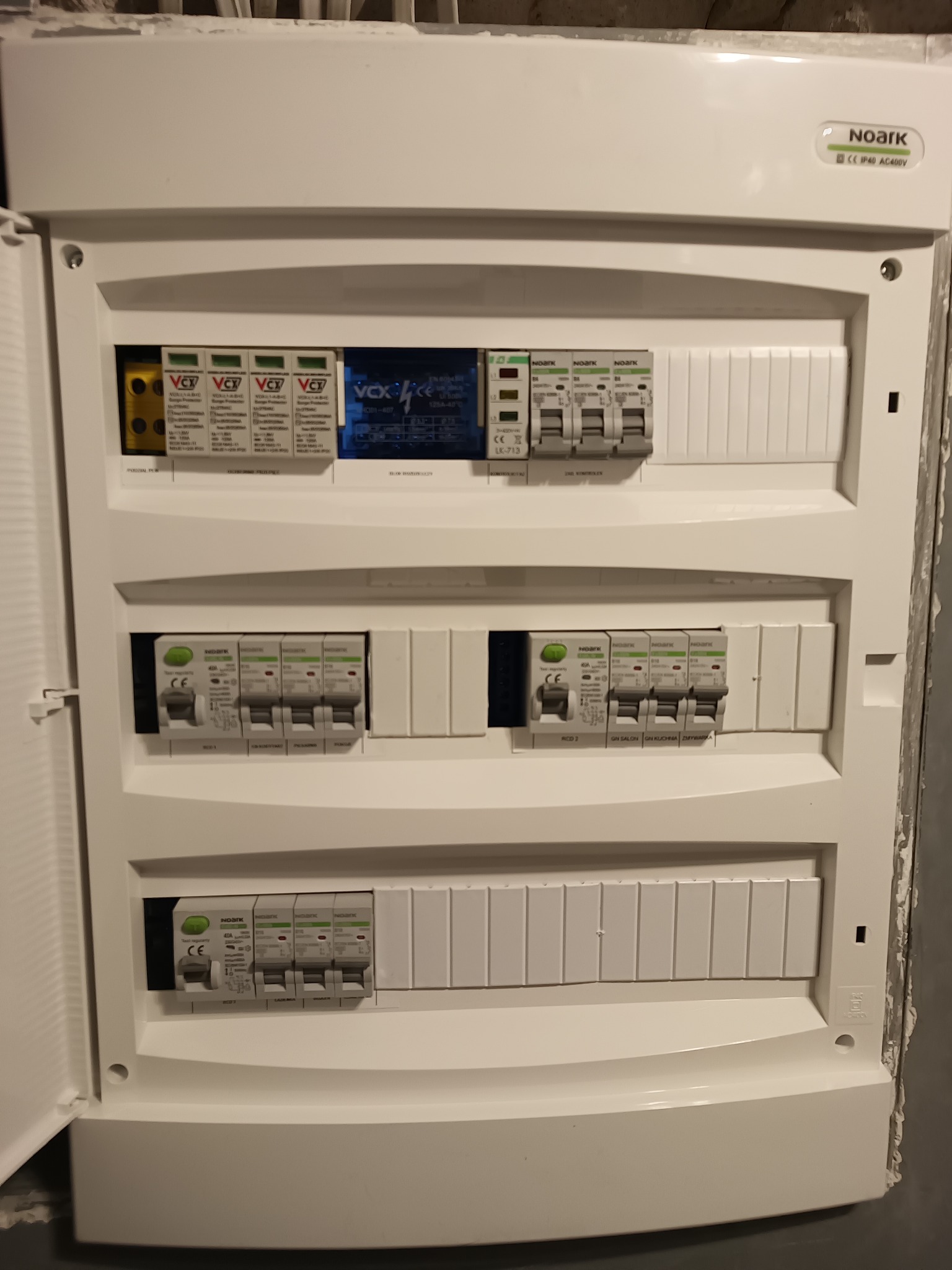 Otwarta skrzynka rozdzielcza z zabezpieczeniami różnicowoprądowymi i ogranicznikami przepięć marki Noark i VCX, widoczne opisy obwodów: salon, kuchnia, zmywarka.