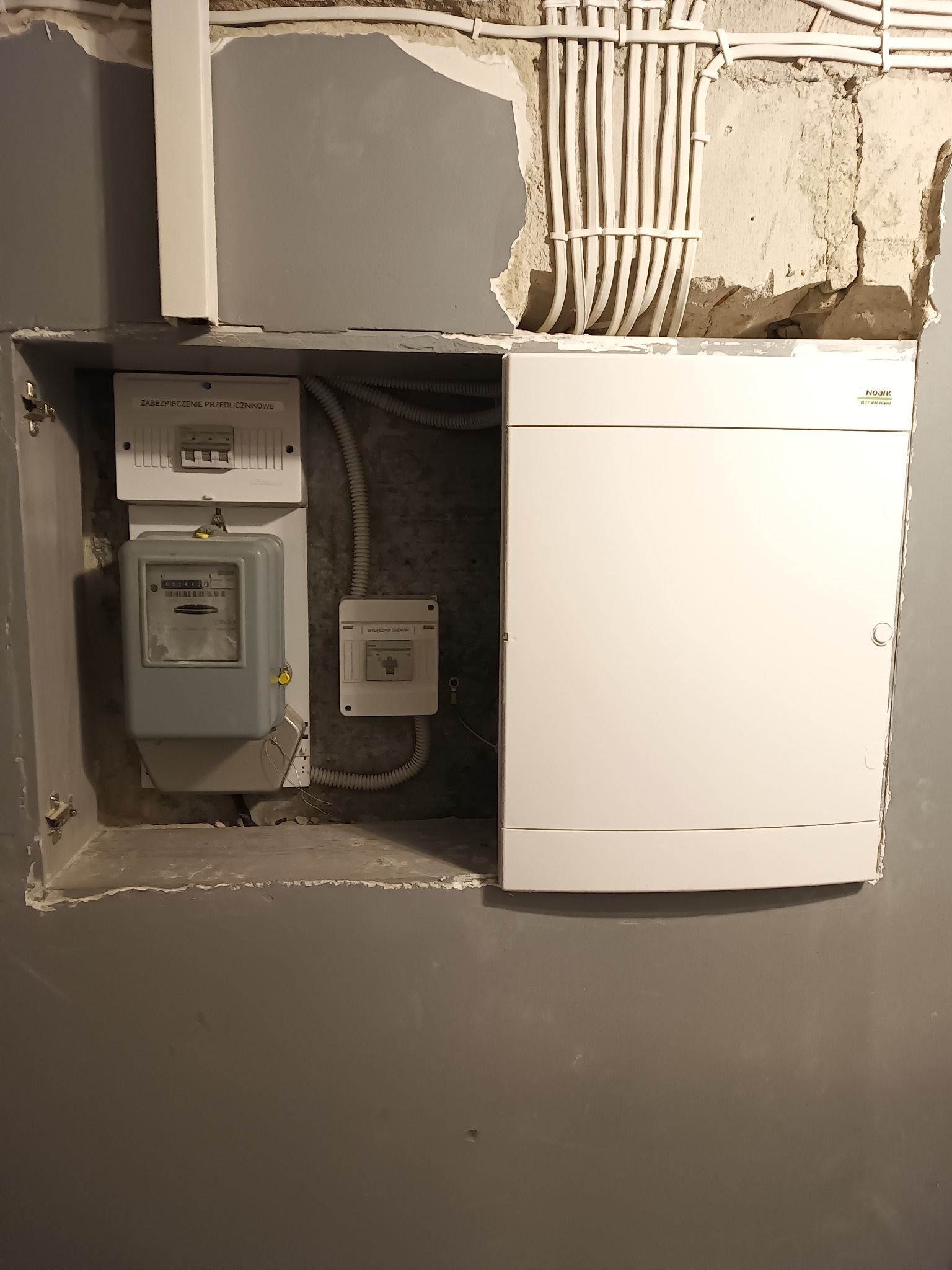 Szara ściana z wnęką, w której umieszczono licznik energii elektrycznej, zabezpieczenie przedlicznikowe i białą skrzynkę rozdzielczą. Nad skrzynką widoczne kable w peszlach.