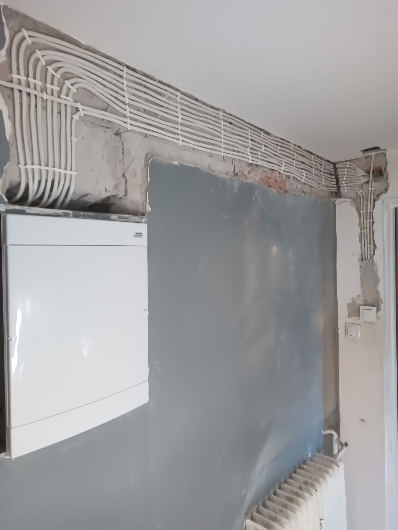 Białe kable elektryczne prowadzone wzdłuż sufitu i ściany w trakcie remontu, widoczna szara ściana, biała skrzynka elektryczna i fragment grzejnika.