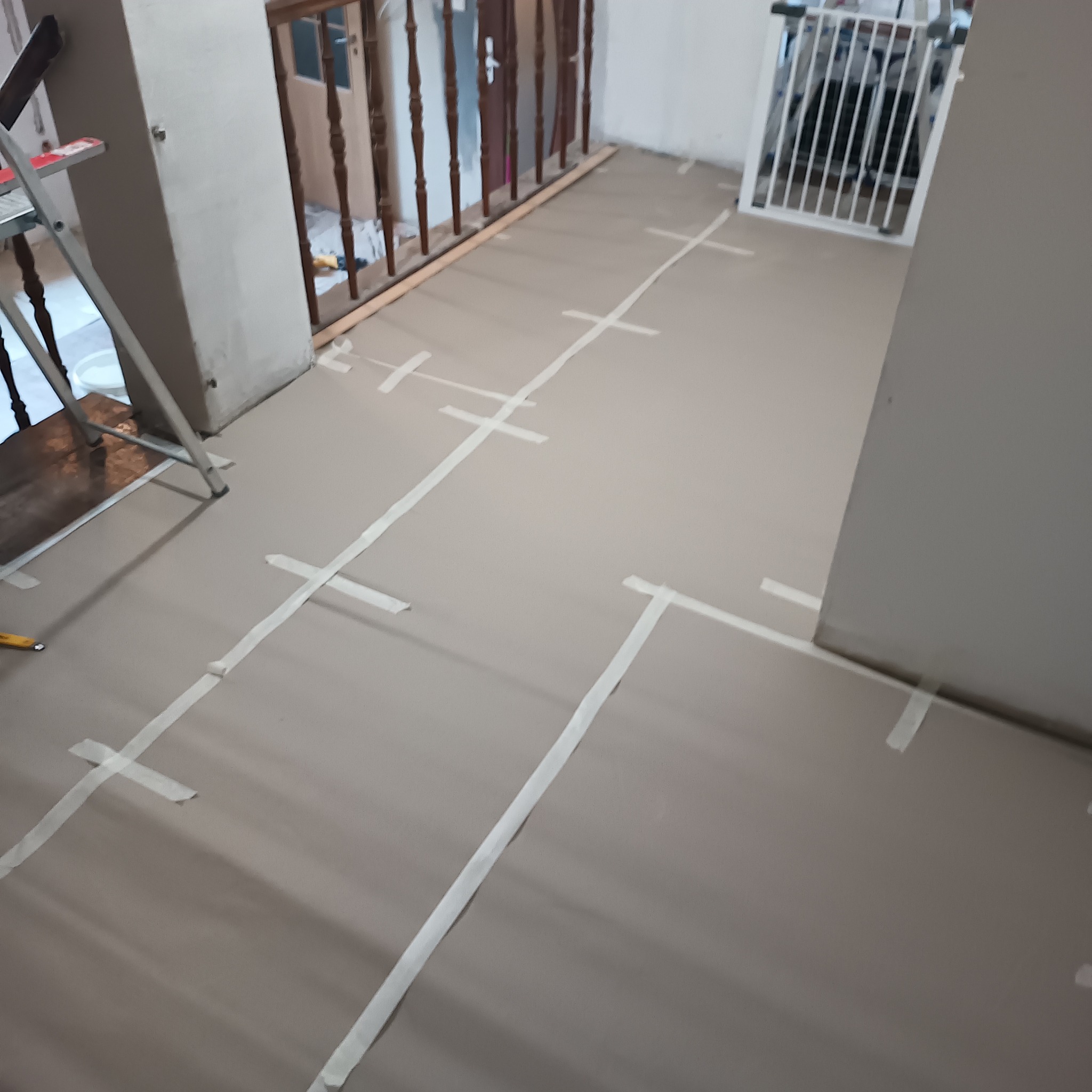 Przygotowanie podłoża pod instalację elektryczną w domu: podłoga z zaznaczonymi liniami taśmą malarską, drabina i barierka zabezpieczająca w tle.