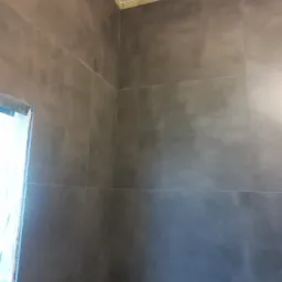 Ściana wyłożona szarymi płytkami imitującymi beton, z widocznym fragmentem otworu drzwiowego zabezpieczonego folią malarską i żółtą taśmą na styku ściany z sufitem, wskazując na prace wykończeniowe...