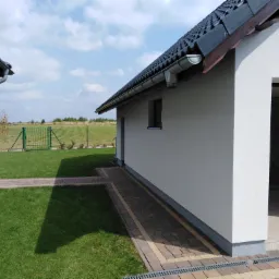 Jasny budynek z garażem i wykończoną elewacją, widoczny z boku, z zadbanym trawnikiem i kostką brukową prowadzącą wzdłuż ściany, na tle zielonego krajobrazu z ogrodzeniem.