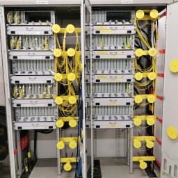 FiberOptic - Otwarta szafa telekomunikacyjna z panelami światłowodowymi, uporządkowanymi żółtymi kablami i oznaczeniami modułów na białych etykietach.